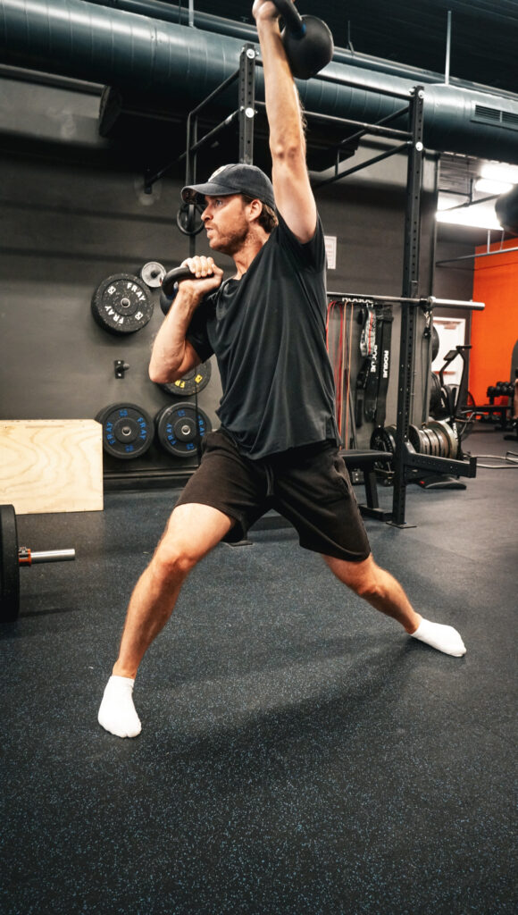 Sander performing a kettlebell exercise at AMPT Fit, standing in a lunge position and lifting the kettlebell overhead. He is focused and demonstrating proper form, reflecting his commitment and progress during his training sessions.